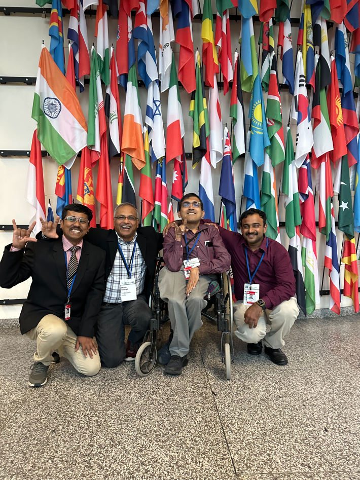 Four men, including one wheelchair users, smiling in front of international flags at an event celebrating disability innovation.