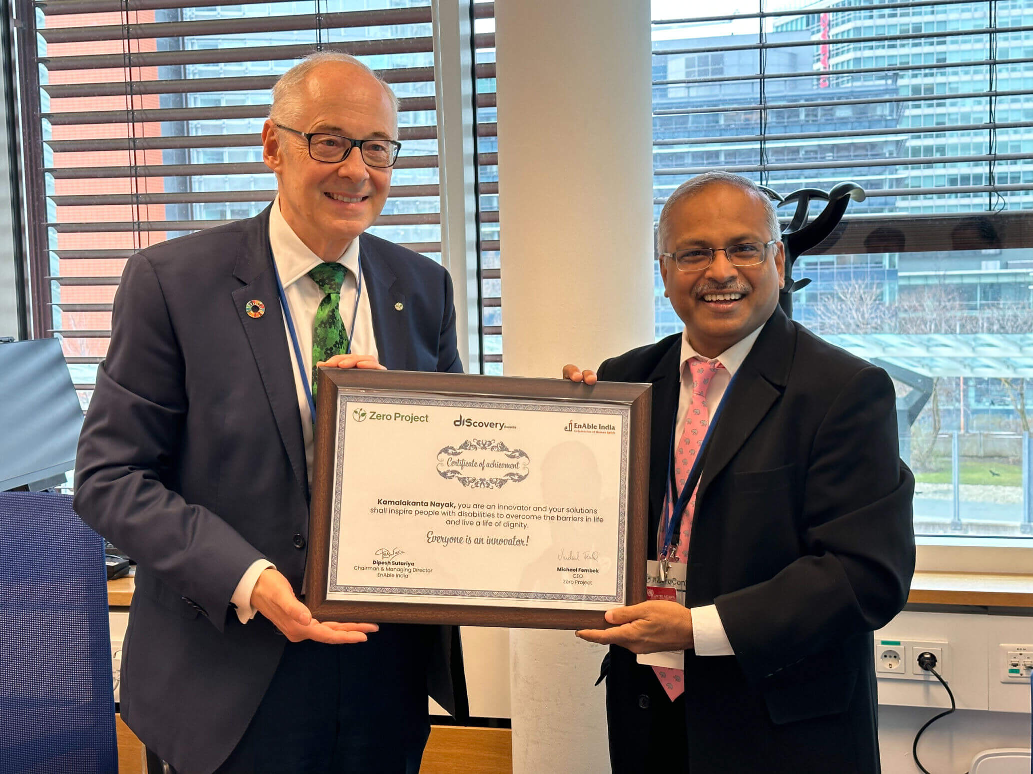 Two men in formal attire standing indoors and smiling while holding a framed certificate of achievement together.