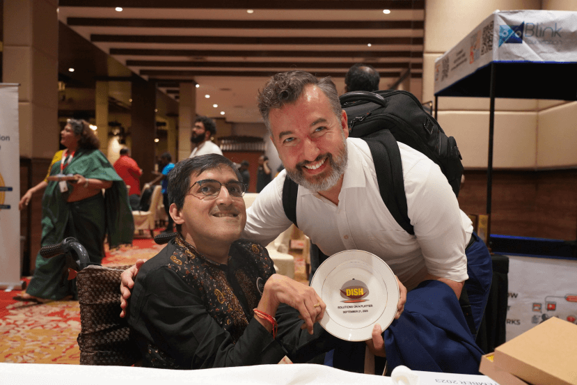 Two smiling men, one using a wheelchair and the other holding a plate that reads 'DISH - Solutions on a Platter', celebrating at an event.