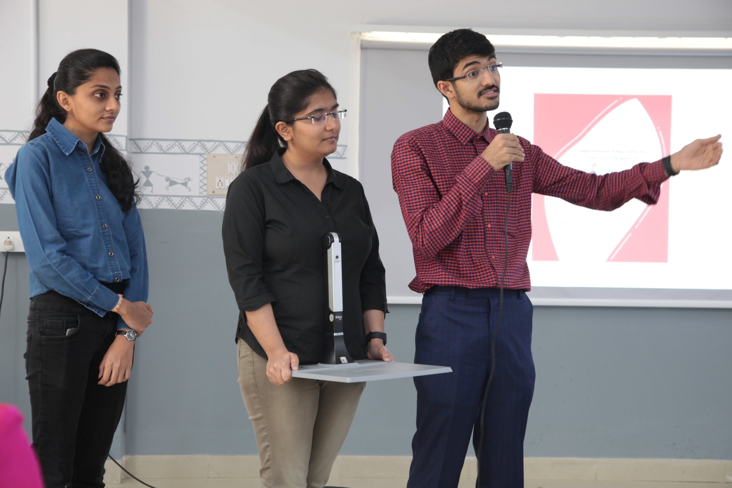 Two young individuals, one speaking into a microphone and the other standing with a tech device on a table, presenting at an EnAble India event.