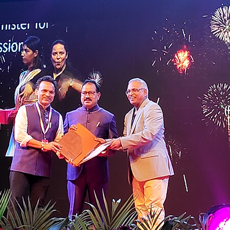 Three men on stage, two receiving a folder from a dignitary, with fireworks visible on the screen behind them.