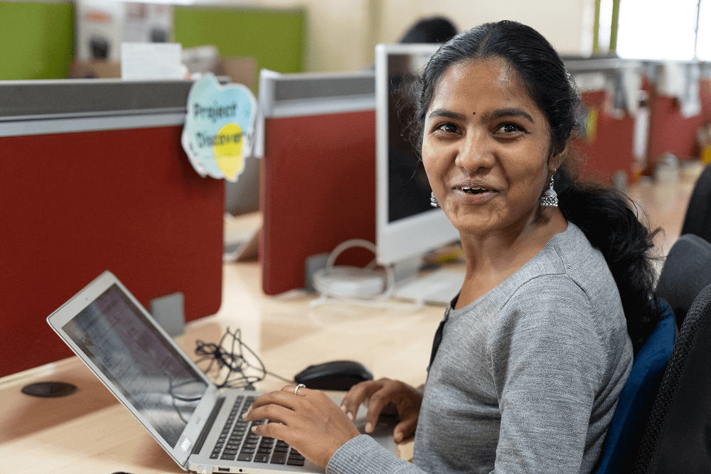 Smiling woman working on a laptop at a cubicle in an office.